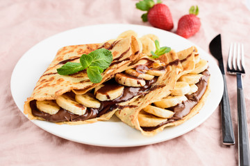 Crepe with chocolate spread and banana on white plate on pink background. Tasty dessert