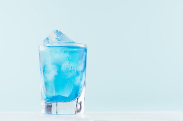 Bright blue tropical alcohol cocktail with curacao liquor, ice cubes in elegant misted shot glass on soft light blue background.