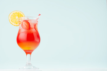 Bright layered red, yellow alcohol cocktail with oranges slice, straw, ice, strawberry  in wineglass on soft light pastel blue background.