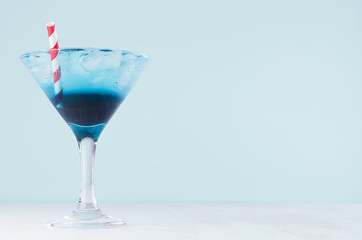 Cold tropical blue drink with ice, red straw in misted wineglass in modern pastel green interior on white wooden table.