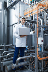 Portrait of man in a white robe and a cap standing in production department of dairy factory with laptop