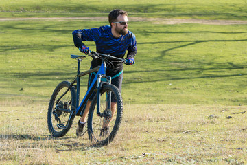 Fototapeta premium Cyclist in shorts and jersey on a modern carbon hardtail bike with an air suspension fork rides off-road on green hills near the forest 