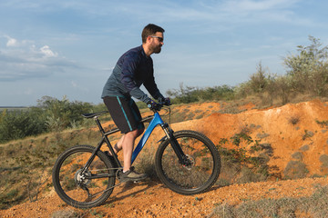 Fototapeta premium Cyclist in shorts and jersey on a modern carbon hardtail bike with an air suspension fork rides off-road on the orange-red hills at sunset evening in summer
