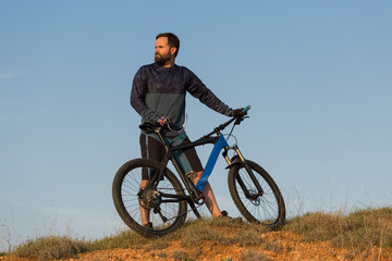 Cyclist in shorts and jersey on a modern carbon hardtail bike with an air suspension fork rides off-road on the orange-red hills at sunset evening in summer