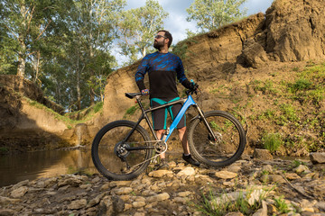 Obraz premium Cyclist in shorts and jersey on a modern carbon hardtail bike with an air suspension fork stands on the rocky shore of a mountain river