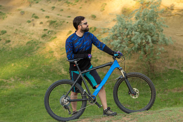 Obraz premium Cyclist in shorts and jersey on a modern carbon hardtail bike with an air suspension fork rides off-road on green hills near the forest 