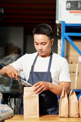 Asian man wearing apron packing freshly roasted coffee beans in craft paper bags while working in roastery house