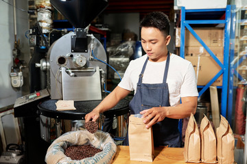 Young Asian factory worker packing roasted coffee beans from bag into paper bag in coffee factory