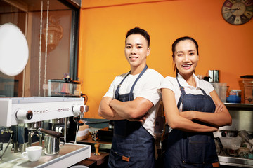 Portrait of young Asian waiter and waitress standing in uniform with arms crossed and smiling at camera near the coffee machine in coffee shop