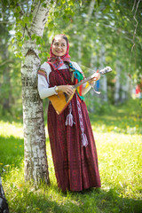 Young thoughtful woman in traditional russian clothes standing in the forest and holding balalaika