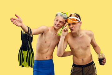 Two young men's half-length portrait isolated on yellow studio background. Smiling friends in caps with drink and flippers. Facial expression, summer, weekend, resort or vacation concept. Trendy
