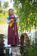 Young smiling woman in traditional russian clothes stands on a small bridge near the lake, and...