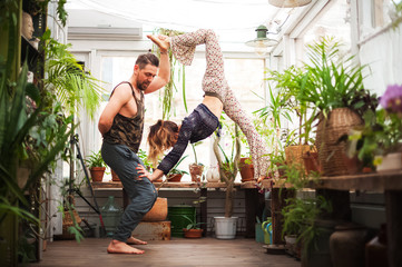 Fototapeta na wymiar A man and women practice yoga asanas on a veranda in a greenhouse with flowers. Pair yoga