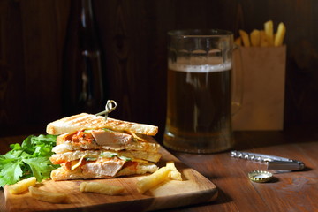 Sandwich with chicken meat, greens and a mug of beer on a dark wooden background. Low key.