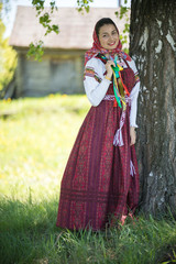 Obraz na płótnie Canvas Young woman in traditional russian clothes standing under a tree and holding her pigtail