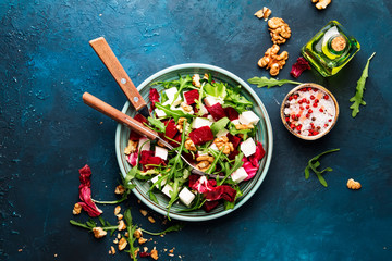 Beet summer salad with arugula, radicchio, soft cheese and walnuts on plate with fork, dressing and spices on blue kitchen table, copy space, top view