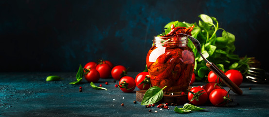 Dried tomatoes in olive oil with green basil and spices in glass jar on blue kitchen table, place for text