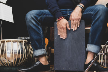 A stylish musician in denim and double monk shoes plays the Cajon, a Peruvian drum used commonly...