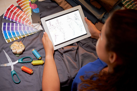 Young designer looking at display of digital tablet and examining the sketches of new collection before sewing while sitting at the table in workshop