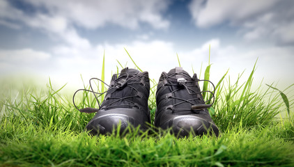 A pair of black trainers, sport shoes on lush green grass with a summer blue sky and white cloud background.