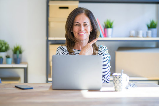 Middle age senior woman sitting at the table at home working using computer laptop cheerful with a smile of face pointing with hand and finger up to the side with happy and natural expression on face
