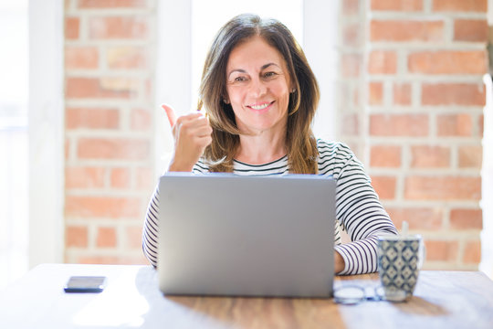 Middle age senior woman sitting at the table at home working using computer laptop pointing and showing with thumb up to the side with happy face smiling