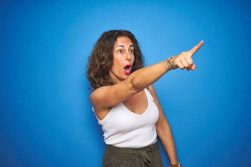 Middle age senior woman standing over blue isolated background Pointing with finger surprised ahead, open mouth amazed expression, something on the front