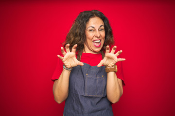 Middle age senior woman wearing apron uniform over red isolated background smiling funny doing claw gesture as cat, aggressive and sexy expression