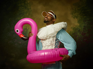 Young man as a medieval grandee or nobleman in a swimring as a pink flamingo on dark studio...