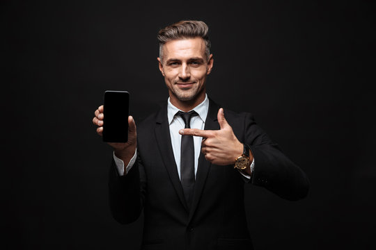 Business man posing isolated over black wall background using mobile phone showing display.