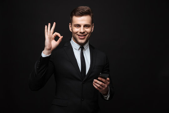 Handsome business man posing isolated over black wall background using mobile phone showing okay gesture.