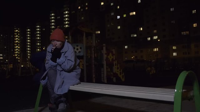 homeless person sits on bench in playground against background of night city. houseless woman is cold and lonely.