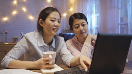 female graphic designer pointing with finger on laptop computer screen talking explain about project. Two asian female friends discussing website sitting at kitchen table in dark night home.