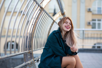 Closeup portrait of pleased blonde girl wearing green coat sitting near the handrail. Space for text