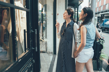 shopping time with friends. young local japanese women standing out clothes store looking beautiful dress of window shop. girl point inside store smiling discussing with sister about fashion by road