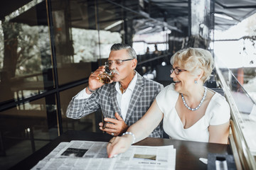 Two beautiful seniors couple drinking wine   at summer terrace ,and reading newspaper at this time