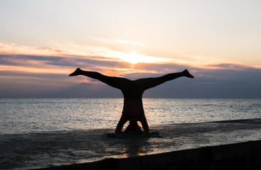 unrecognizable woman with beautiful body doing yoga headstand at sunrise on the sea, silhouette of yoga poses