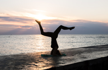 unrecognizable woman with beautiful body doing yoga headstand at sunrise on the sea, silhouette of yoga poses