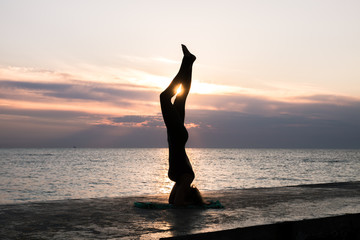 unrecognizable woman with beautiful body doing yoga headstand at sunrise on the sea, silhouette of yoga poses