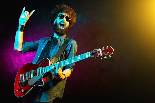 Young hipster man with curly hair with red guitar show rock gesture in neon lights. Rock musician is playing electrical guitar.