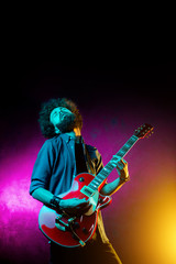 Young hipster man with curly hair with red guitar in neon lights. Rock musician is playing electrical guitar.