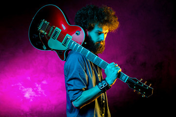 Obraz premium Portrait of hipster man with curly hair with red guitar in neon lights. Rock musician is playing electrical guitar.