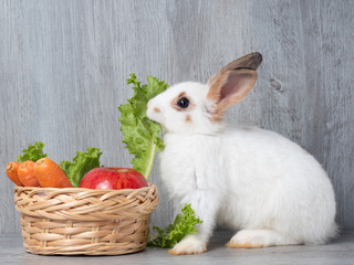 White cute rabbit  eating green lettuce carrot and apple in the wooden basket and wooden gray background. Rabbits like to eat vegetables but should not give too much. © arlee