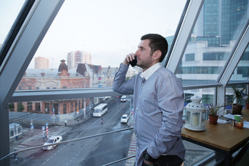 A young businessman wearing a shirt with stubble on his face is talking on a mobile phone against the background of a panoramic window on a high floor overlooking the street, buildings and cars.