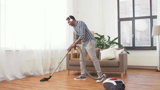 household and cleaning concept - indian man in headphones with vacuum cleaner at home