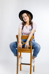 Young beautiful girl sitting on wooden chair in stylish black hat on white background. Emotions and gestures.