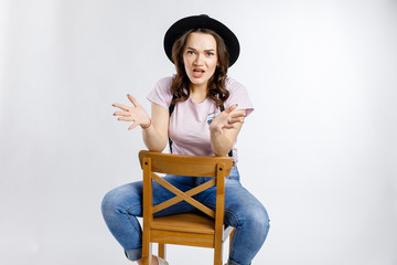 Young beautiful girl sitting on wooden chair in stylish black hat on white background. Emotions and gestures.