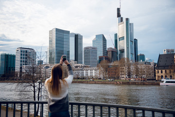 Girl with long straight hair photographs skyscrapers near river in Frankfurt am Main. Focus on the city