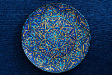 Decorative ceramic plate with blue and golden colors, painted plate on the background of blue fabric, closeup. Decorative porcelain plate painted with acrylic paints, handwork