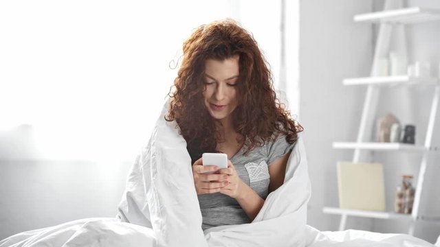 beautiful girl covered in blanket using smartphone and smiling in bed in morning